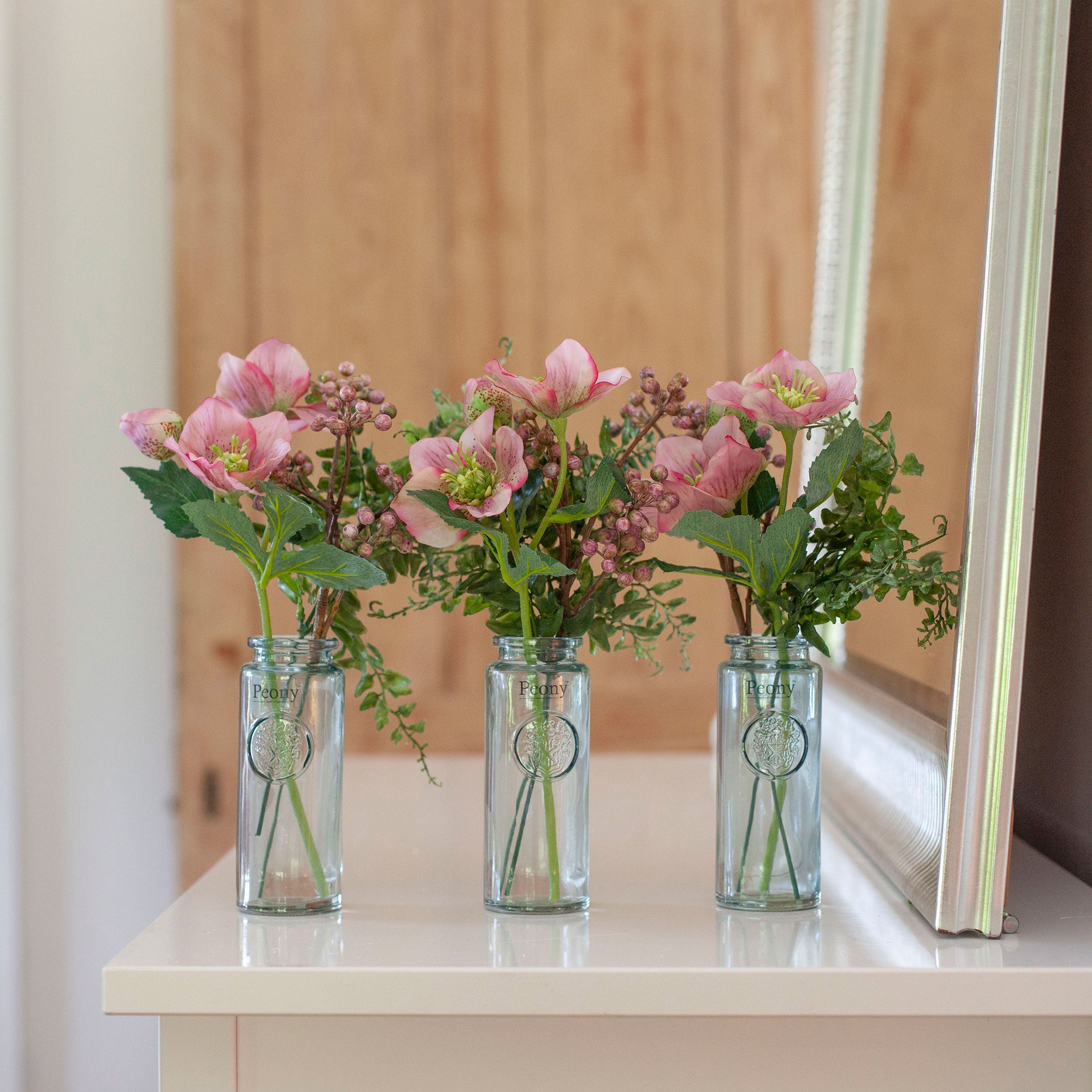 Set of three artificial hellebore and berry arrangements in  pink, arranged in individual glass bottle vases and styled on a console table.