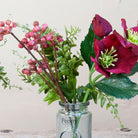 Close-up of an artificial burgundy hellebore and berry arrangement with green foliage in a glass bottle vase.