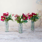 Set of three artificial burgundy hellebore and berry arrangements with green foliage in individual glass bottle vases.