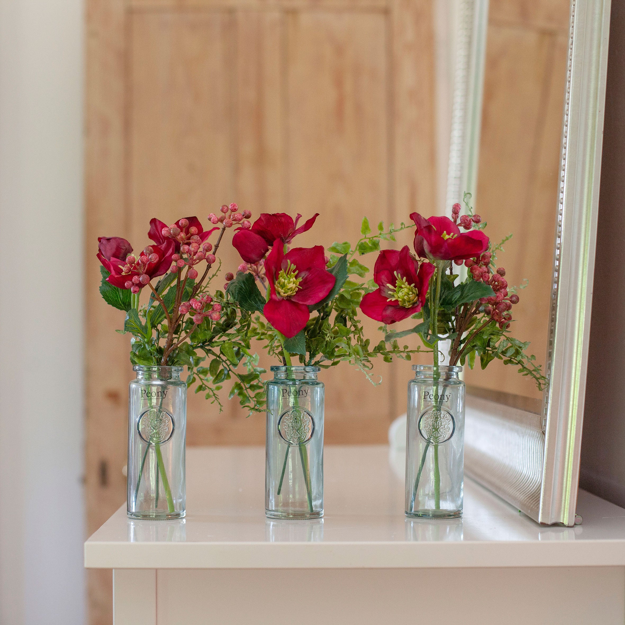 Set of three artificial hellebore and berry arrangements in  beuaty, arranged in individual glass bottle vases and styled on a console table.