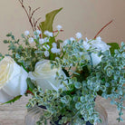 Close-up of artificial white roses, mini eucalyptus and gypsophila set in a clear optic glass jar vase.