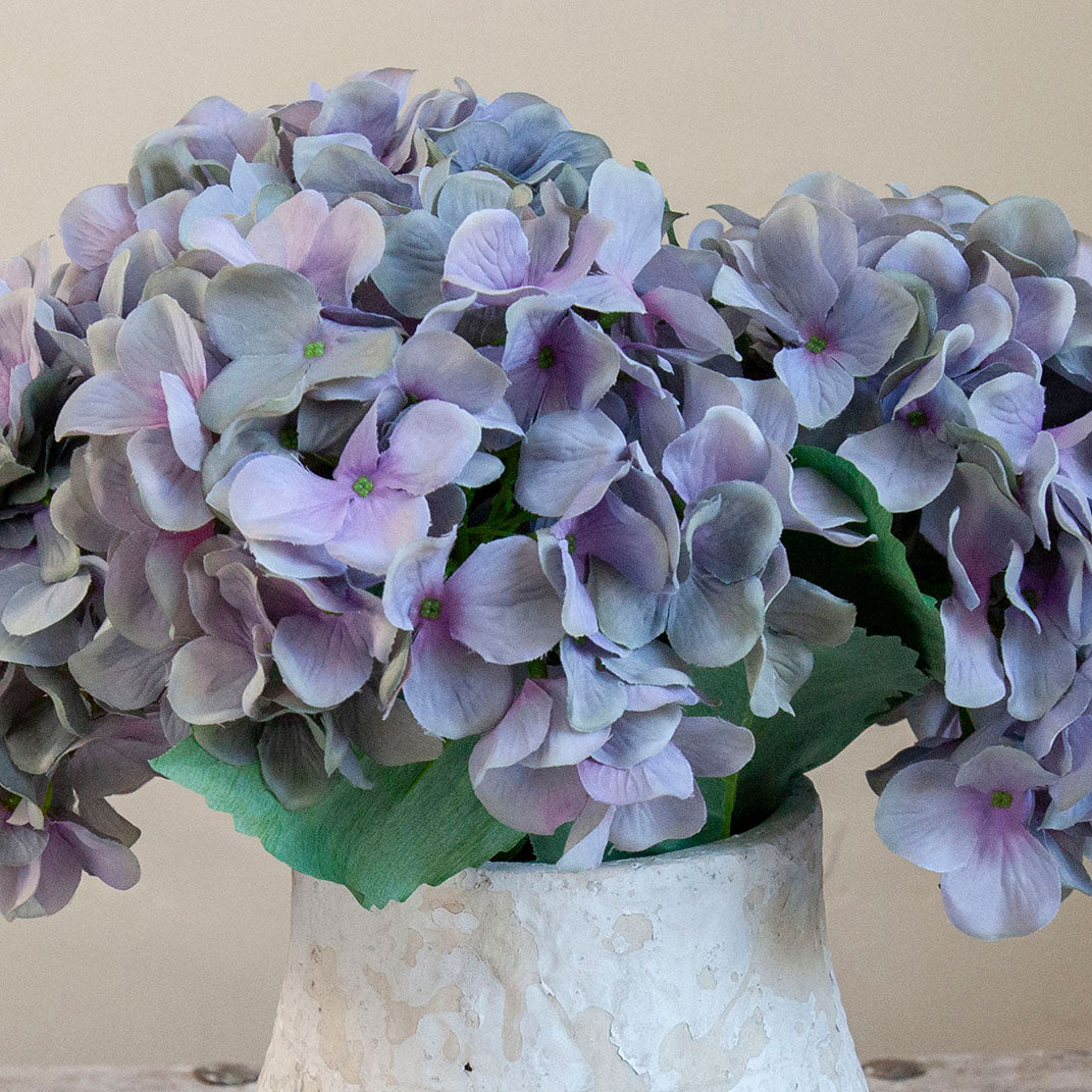 Close-up of artificial hydrangea blooms in soft blue, lilac and grey tones in a rustic textured white pot