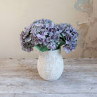 Artificial hydrangea arrangement in soft blue, lilac and grey tones in a rustic textured white pot.
