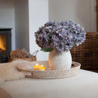 Artificial hydrangea arrangement in soft blue, lilac and grey tones in a rustic textured white pot, styled on a woven tray in a cosy living room.