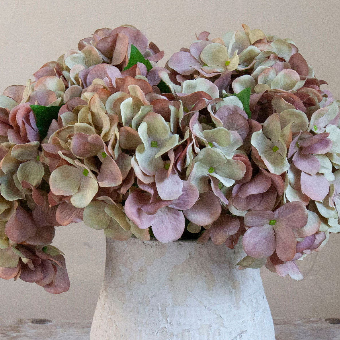 Close-up of artificial hydrangea blooms in soft blush, mauve and green tones in a rustic textured white pot.