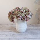 Artificial hydrangea arrangement in soft blush, mauve and green tones in a rustic textured white pot.