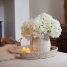 Artificial white hydrangea arrangement in a rustic textured pot, styled on a woven tray in a cosy living room.