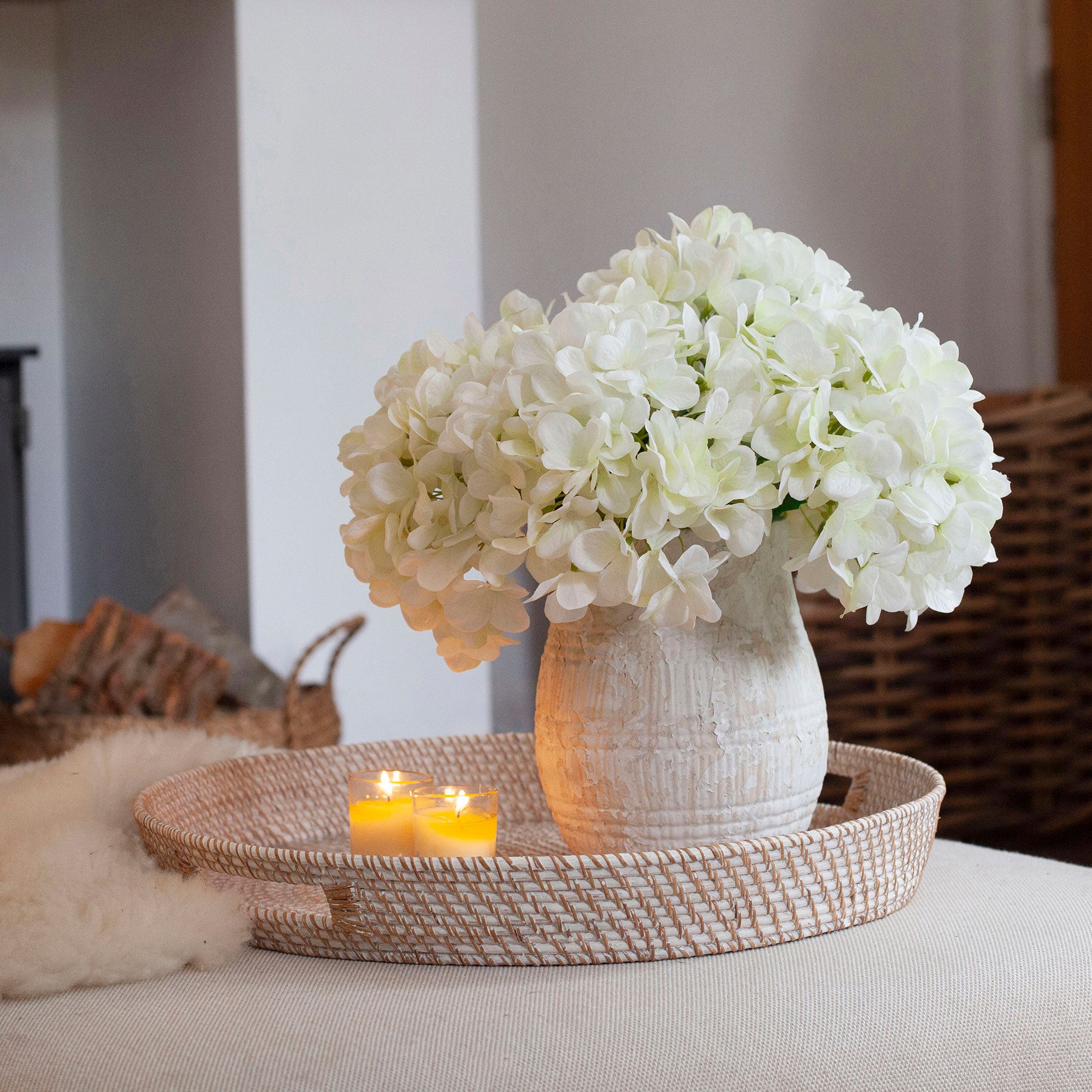 Artificial white hydrangea arrangement in a rustic textured pot, styled on a woven tray in a cosy living room.