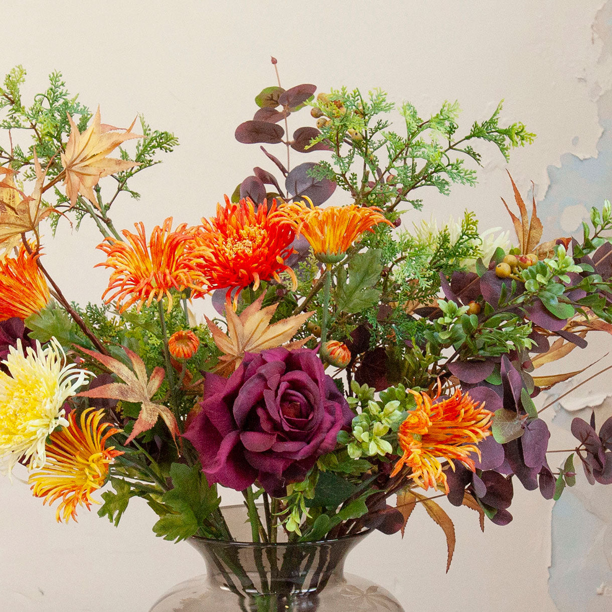 Close up of artificial chrysanthemum and autumn foliage bouquet in rich orange, burgundy and green tones, displayed in a smoked glass vase.
