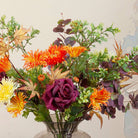 Close up of artificial chrysanthemum and autumn foliage bouquet in rich orange, burgundy and green tones, displayed in a smoked glass vase.