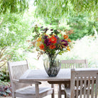 Artificial chrysanthemum and autumn foliage bouquet in rich orange, burgundy and green tones, displayed in a smoked glass vase on an outdoor garden table.