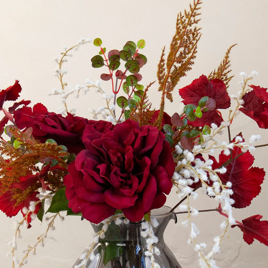 Close up of Artificial Rose, Grape Leaf & Acacia Arrangement with rich burgundy roses, red grape foliage, white acacia and warm autumnal accents displayed in a smoked pumpkin glass vase