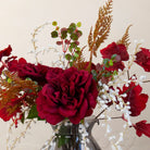 Close up of Artificial Rose, Grape Leaf & Acacia Arrangement with rich burgundy roses, red grape foliage, white acacia and warm autumnal accents displayed in a smoked pumpkin glass vase