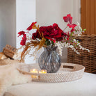 Lifestyle shot of Artificial Rose, Grape Leaf & Acacia Arrangement with rich burgundy roses, red grape foliage, white acacia and warm autumnal accents in a smoked pumpkin glass vase