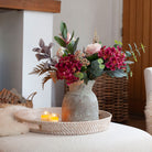 Artificial rose and hydrangea arrangement in soft pink and rich burgundy tones with eucalyptus and autumn foliage, styled in a rustic vase in a cosy living room.