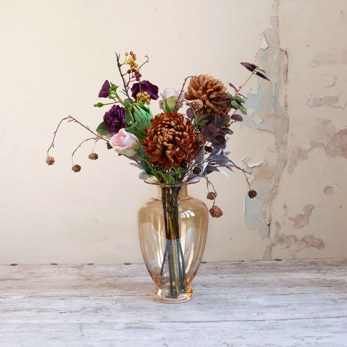 Artificial chrysanthemum, rose and foliage bouquet in warm autumn tones, displayed in an amber glass vase.