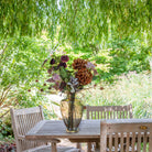 Artificial chrysanthemum, rose and foliage bouquet in warm autumn tones, displayed in an amber glass vase on an outdoor garden table.