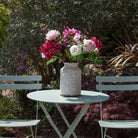 Artificial hydrangea, dahlia and peony bouquet in pink, burgundy and white tones, displayed in a white textured vase on an outdoor garden table.