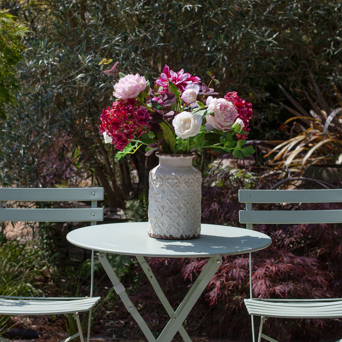 Artificial hydrangea, dahlia and peony bouquet in pink, burgundy and white tones, displayed in a white textured vase on an outdoor garden table.