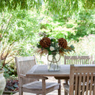 Artificial chrysanthemum, rose and astilbe bouquet in warm autumn tones, displayed in a clear glass rope vase on an outdoor garden table