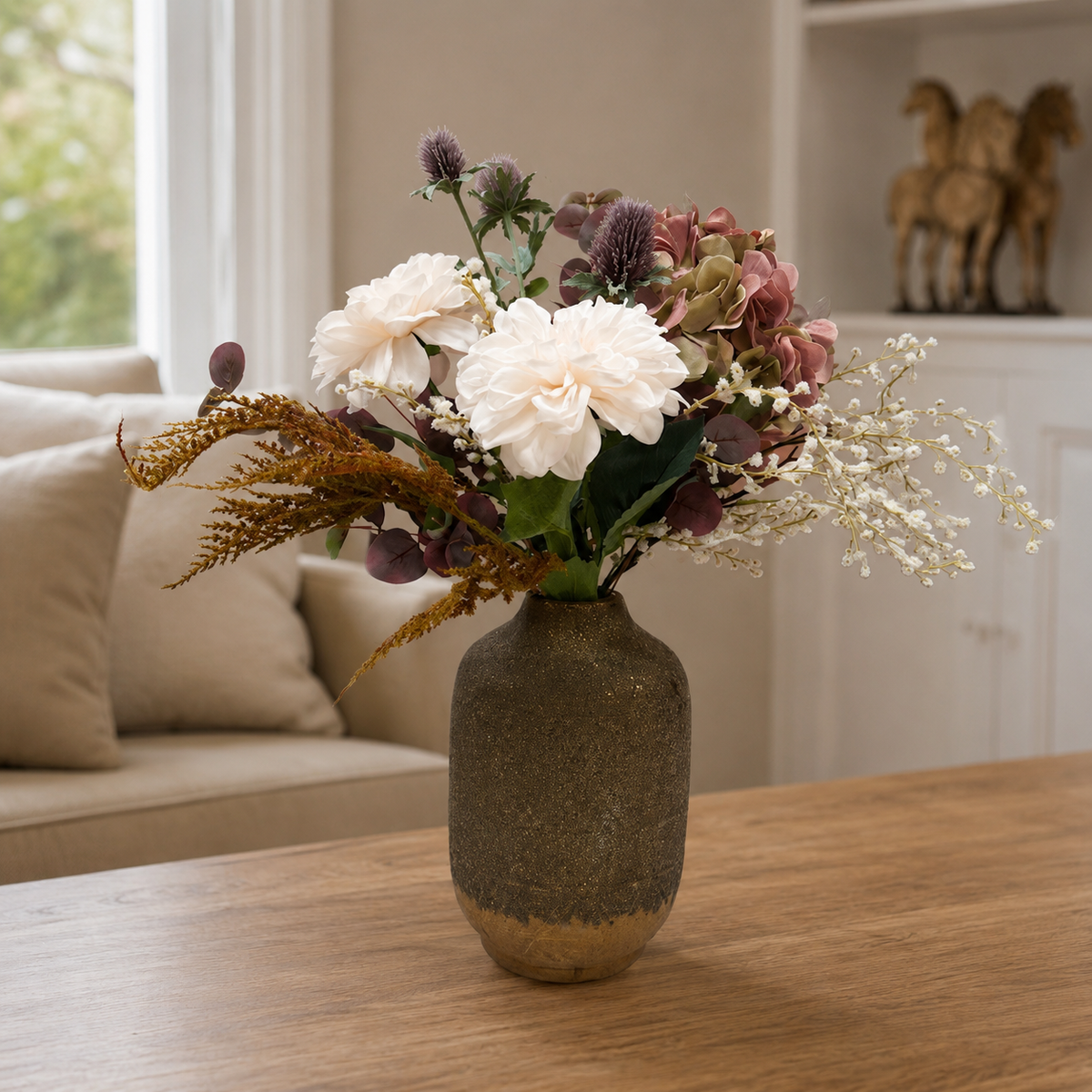 Artificial dahlia, hydrangea and thistle bouquet in soft white, blush and muted autumn tones, styled in a textured bronze vase on a wooden table.