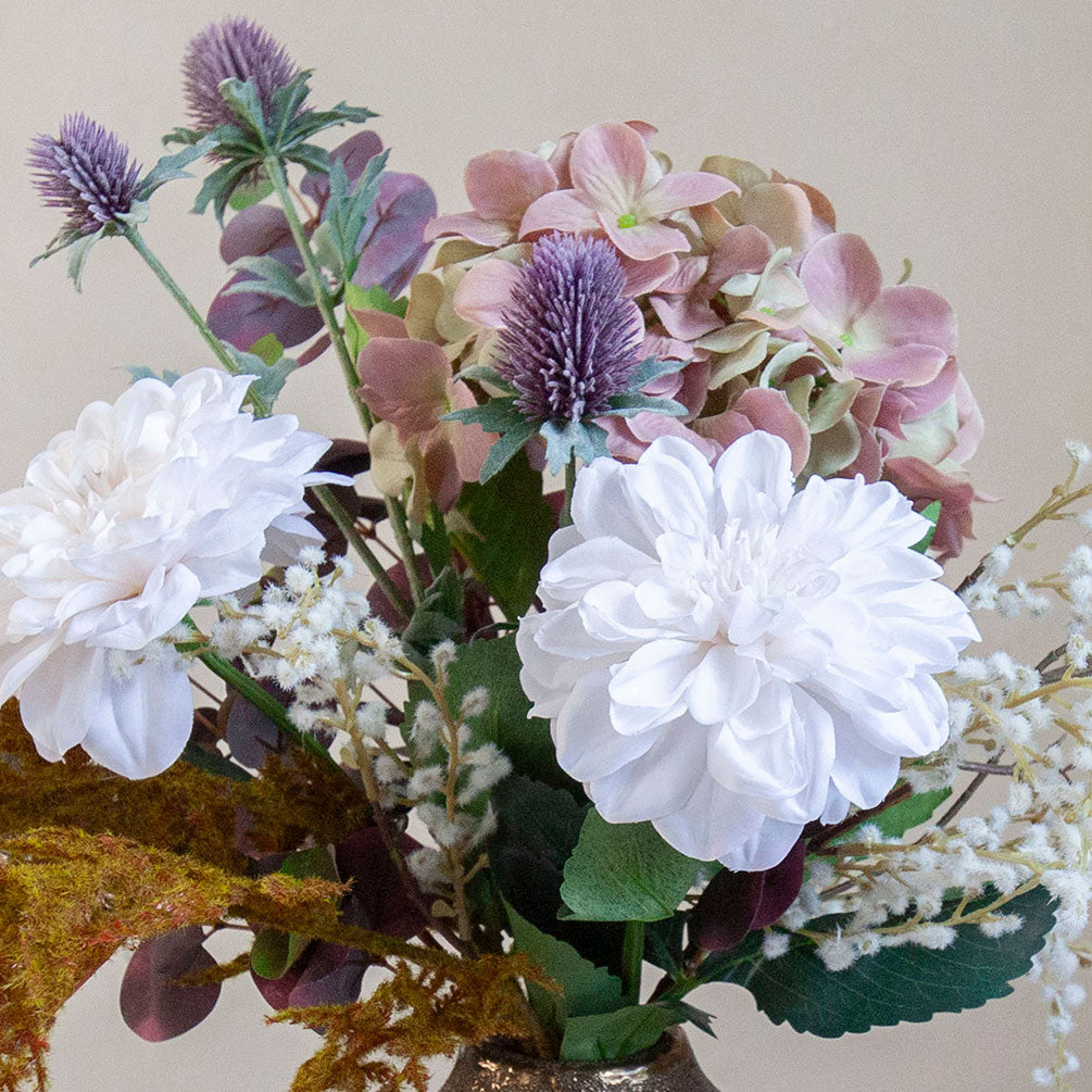 Close up of artificial dahlia, hydrangea and thistle bouquet in soft white, blush and muted autumn tones, displayed in a textured bronze vase.