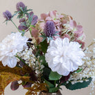 Close up of artificial dahlia, hydrangea and thistle bouquet in soft white, blush and muted autumn tones, displayed in a textured bronze vase.