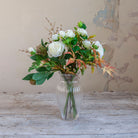 Artificial rose, ranunculus and autumn foliage bouquet with white flowers and warm seasonal leaves, displayed in a clear glass vase.