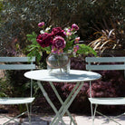 Artificial roses, hellebores and foliage in rich burgundy and mauve tones, displayed in a clear glass rope lantern vase on an outdoor garden table.