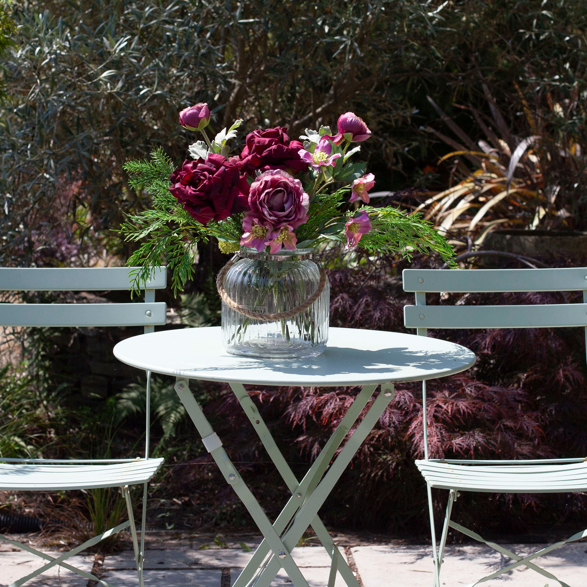 Artificial roses, hellebores and foliage in rich burgundy and mauve tones, displayed in a clear glass rope lantern vase on an outdoor garden table.