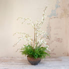 Artificial white dancing orchids with fern foliage arranged in a bronze footed bowl.