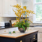 Artificial yellow dancing orchids with fern foliage arranged in a bronze footed bowl, styled on a kitchen island.
