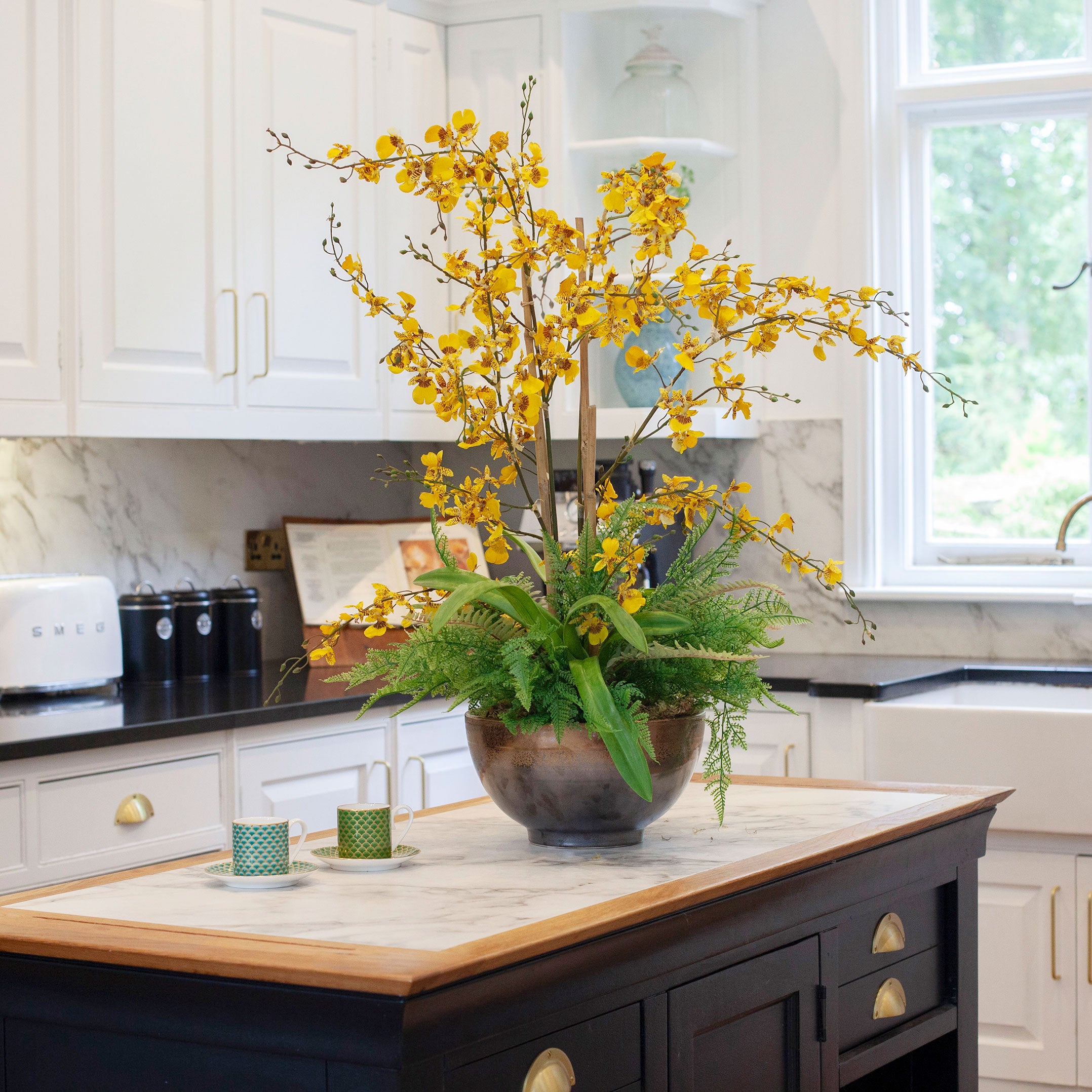 Artificial yellow dancing orchids with fern foliage arranged in a bronze footed bowl, styled on a kitchen island.