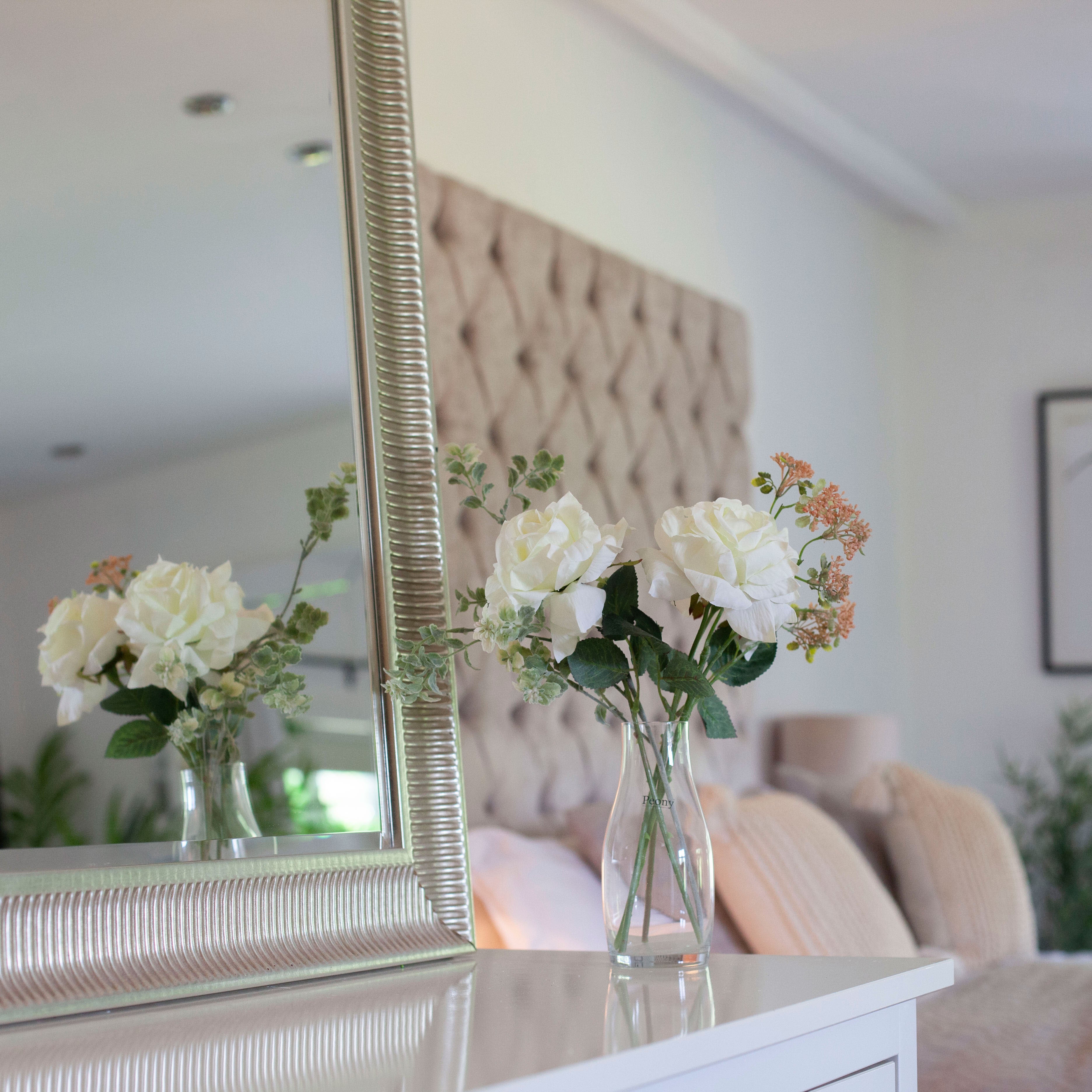 Artificial white roses and patrinia set in a clear glass milk bottle vase, styled on a bedroom dressing table.