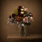 Artificial garden roses, burgundy orchids and bronze chrysanthemums with silvery foliage and dark berries, arranged in a clear glass vase.