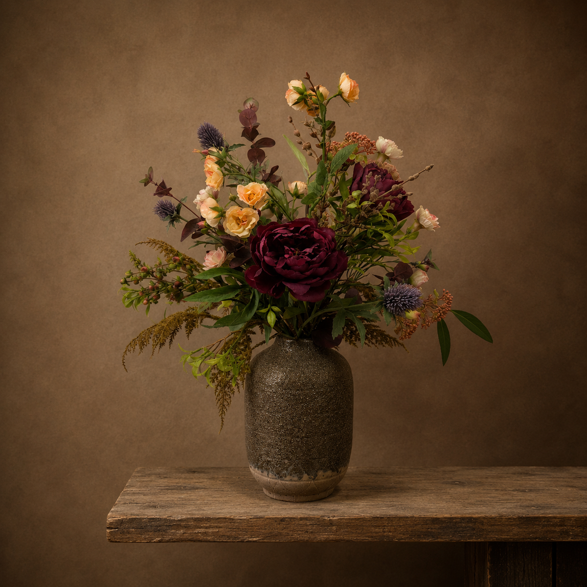 Artificial burgundy peony, soft peach spray roses and purple thistles with autumn foliage, arranged in a tall rustic textured vase.