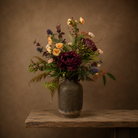 Artificial burgundy peony, soft peach spray roses and purple thistles with autumn foliage, arranged in a tall rustic textured vase.