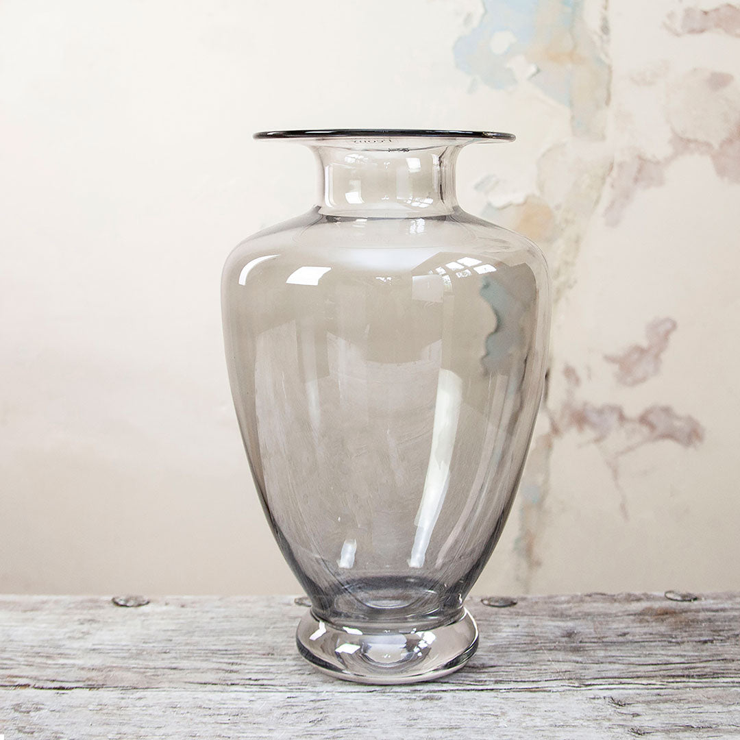 A clear, smoked glass Grecian vase with a lustre finish, placed on a wooden surface against a textured wall.