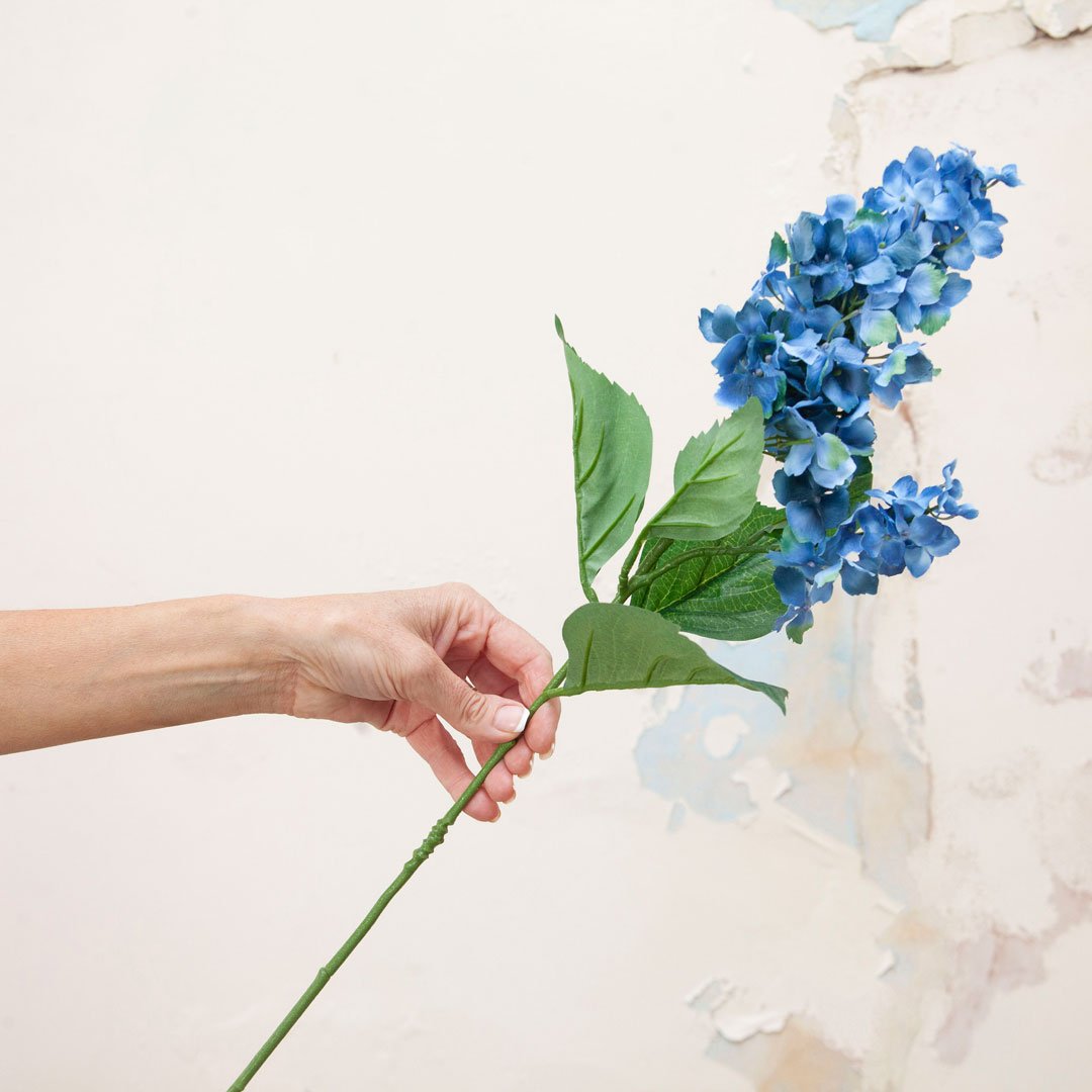 Artificial blue conehead hydrangea stem held in hand, featuring elongated cone-shaped flower heads and green foliage.
