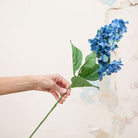 Artificial blue conehead hydrangea stem held in hand, featuring elongated cone-shaped flower heads and green foliage.