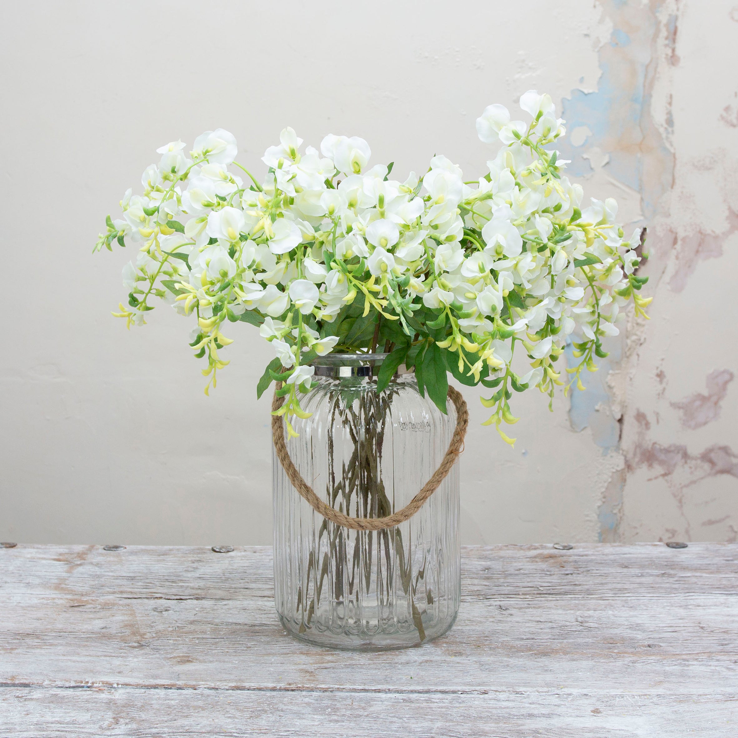 Artificial white wisteria stem displayed in a vase, showcasing trailing clusters of delicate white flowers and natural green leaves.