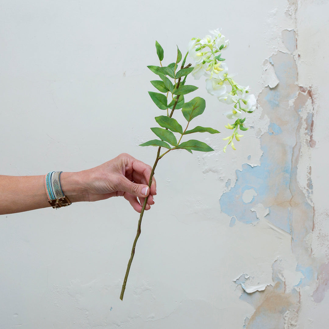 Artificial white wisteria stem being held in hand, featuring cascading white blooms with soft green buds and realistic foliage.