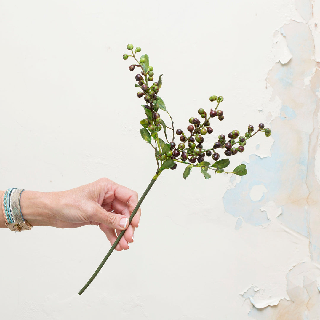 Artificial green and deep berry-toned berry stem being held in hand, featuring branching sprays of small round berries in shades of olive green, plum and deep burgundy, with realistic foliage 
