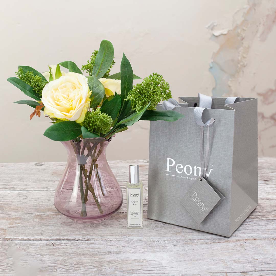 Artificial rose and skimmia arrangement displayed in a modern Prisma vase, presented with a gift bag and a 25ml rose fragrance bottle.
